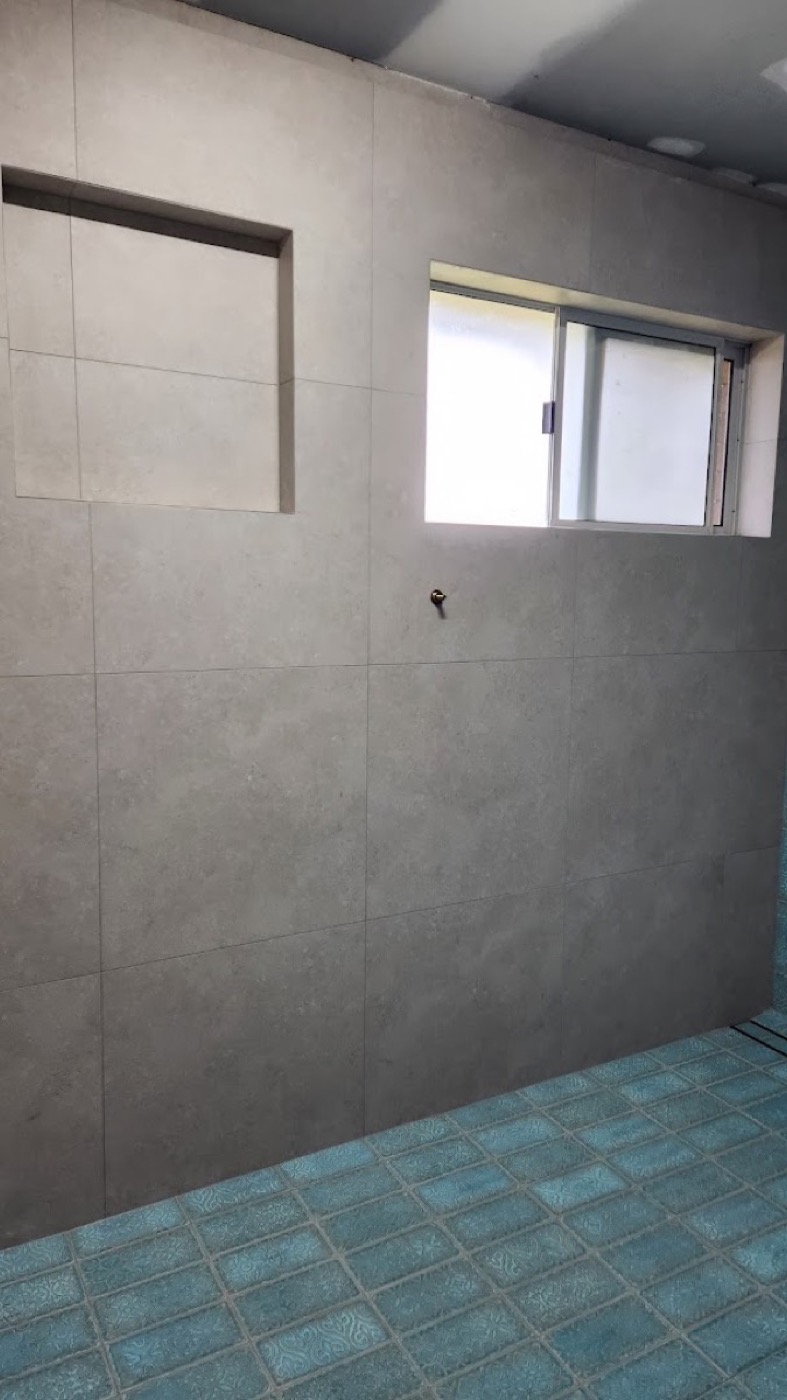 A part-tiled wall in large grey format tiles beside a window opening, mid job